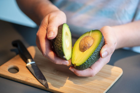 Close-up of hands cutting a ripe avocado in half with a knife on a wooden cutting board in a kitchen. Visible pit, natural light and fresh ingredients create an authentic home cooking scene focused on healthy food preparation and everyday lifestyle.の写真素材