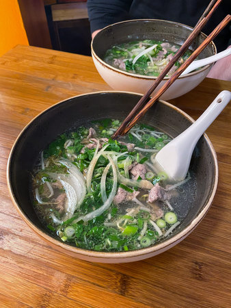 Traditional Vietnamese Pho Bo beef noodle soup served in a ceramic bowl with rice noodles, sliced beef, fresh herbs and green onions. Hot clear broth with aromatic ingredients, photographed on a wooden table in a casual restaurant setting. Popular Asian comfort food and authentic street food cuisine.の写真素材