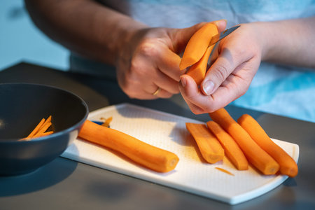 Close-up of hands cutting fresh carrots with a knife on a white cutting board. Several peeled carrots lie next to a bowl in a kitchen setting. Natural food preparation scene showing healthy vegetables and everyday home cooking.の写真素材