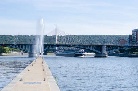 LiÃ¨ge, Belgium - August 22 2019: A boat on the Meuse river in front of the FragnÃ©e bridge and a fountain asideのeditorial素材