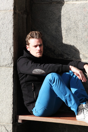 Young man sitting down against a wall of stone, wearing a black hoodie and blue jeans with sunglasses.の写真素材