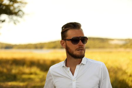 A man wearing a white shirt and dark sunglasses, standing against a large yellow field looking away from camera. A great sunny summer day.の写真素材