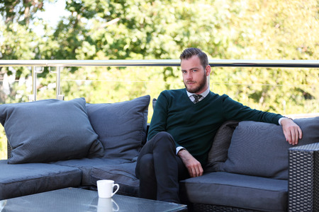 A handsome man in a shirt, tie and sweater sitting down outside on a summer day holding a mug. Relaxed and laid back.の写真素材