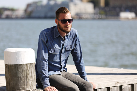 A lonely man sitting down outdoors wearing a jeans shirt and sunglasses, sitting close to the water in a harbor.の写真素材