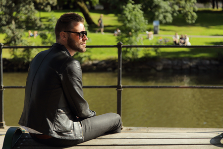 An from the side angle photo of a man sitting on a bench wearing a black leather jacket, with a river and grass behind him. Sunny summer day.の写真素材