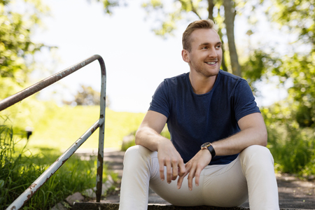 Handsome blonde man in a blue shirt and beige pants sitting down on steps outdoors on a sunny summer day with a big smile on his face.の写真素材