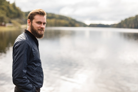 Laid back man with beard standing against a big lake looking at camera outdoors on a sunny day.の写真素材