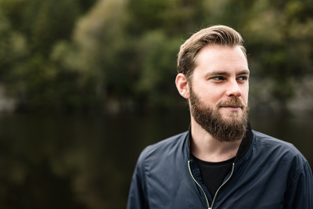 One bearded man wearing a blue jacket, standing in front of a lake and trees on a moody day.の写真素材