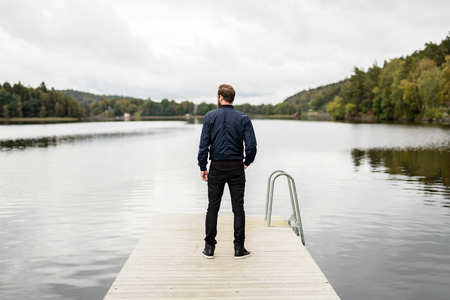 A pensive bearded man wearing a blue jacket and jeans, standing outside with a big lake with surrounding woods.の写真素材