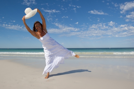 An attractive looking mature woman in her fifties, wearing a white summer dress, is dancing and jumping at a tropical beach with a wide rimmed white hat, with ocean, horizon and blue sky as background and copy space の写真素材