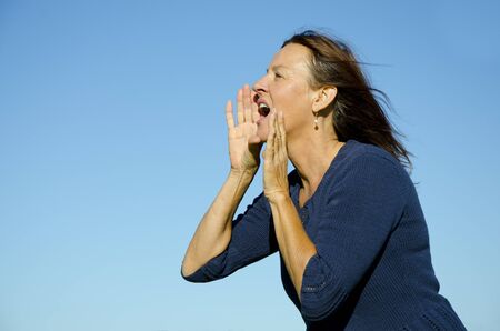 Attractive mature woman with brown hair shouts out loud at a sunny remote outdoor scenery with blue...