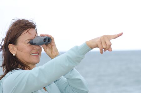 Attractive looking mature woman looking through binoculars and point with one arm, happy, optimistic, confident  Isolated with ocean and white sky as background and copy space の写真素材