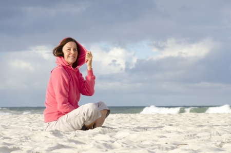 Lonely but happy woman contemplating, dreaming, keeping herself warm outdoor, isolated with ocean and dark storm sky as background and copy space の写真素材
