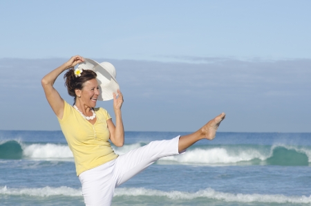Happy healthy retired woman at the beach, stretching one leg in the air, cheerful laughing, isolated with ocean and sky as background and copy space の写真素材