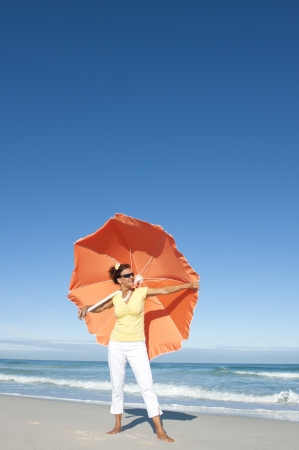 Beautiful looking senior woman standing happy and cheeful with big orange umbrella at beach enjoying retirement, isolated with ocean and blue sky as background and copy space の写真素材