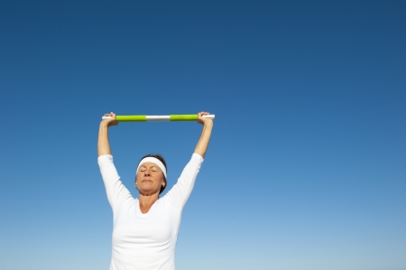 Attractive and active senior woman relaxed standing with weight over her head doing healthy exercise, isolated with blue sky as background and copy space.の写真素材