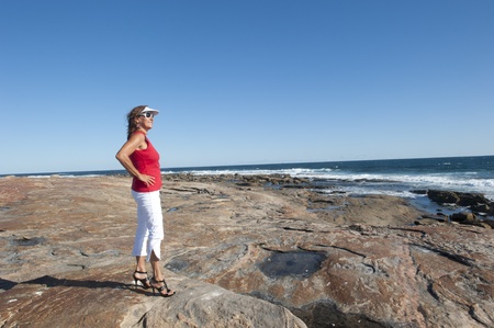 Sexy middle aged woman standing at the ocean wearing red top, white pants and high heel shoes, isolated with happy relaxed smile and sea and blue sky as background and copy space.の写真素材