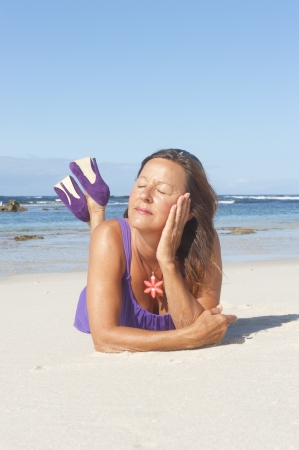 Pretty looking mature woman in sexy purple dress and high heel shoes at the beach, with ocean and blue sky as background and copy space.の写真素材