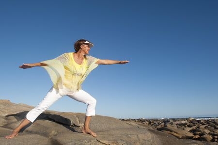 A beautiful looking mature woman standing confident and happy on a rock, isolated with ocean and clear blue sky as background and copy space.の写真素材