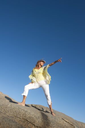 A beautiful looking mature woman standing confident and happy on a rock, isolated with clear blue sky as background and copy space.の写真素材
