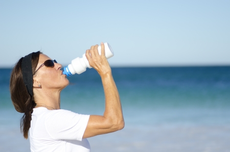 Attractive and active senior woman drinking refreshing water after exercising, isolated with ocean and blue sky as background and copy space.の写真素材