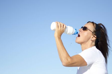 Attractive and active mature woman drinking refreshing water after exercising, isolated with blue sky as background and copy space.の写真素材