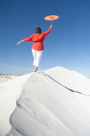A mature woman is balancing on a sheer white sand dune in the desert, wearing a red blouse and a colour-matching umbrellaの写真素材