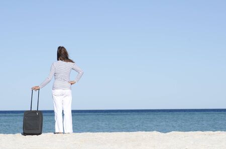 Attractive lady on holiday standing with suitcase alone at a beach overlooking the sea, isolated with ocean and blue sky as background and copy space.の写真素材