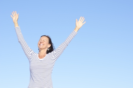 Beautiful happy looking mature woman in cheerful, positive, optimistic pose with arms up, isolated with blue sky as background and copy space.の写真素材