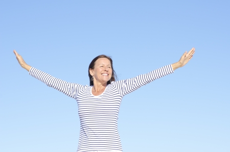 Beautiful happy looking mature woman in cheerful, positive, optimistic pose with arms up, isolated with blue sky as background and copy space.の写真素材