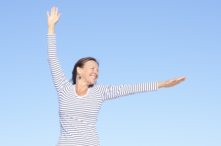 Beautiful happy looking mature woman in cheerful, positive, optimistic pose with arms up, isolated with blue sky as background and copy space.の写真素材