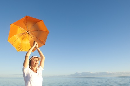 Confident woman in white shirt and pants standing with orange umbrella over shoulder at peaceful ocean at Shark Bay, Western Australiaの写真素材