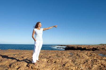 Attractive and confident mature woman standing on top of cliff overlooking ocean, isolated with blue sky as background and copy space.の写真素材