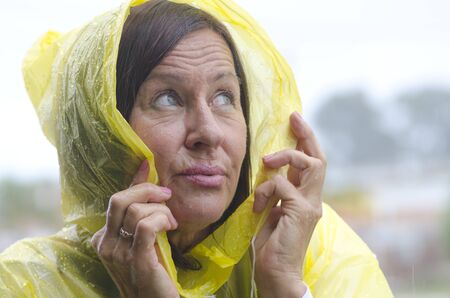 Portrait mature woman standing in the rain, wearing yellow raincoat, isolated with grey sky as...