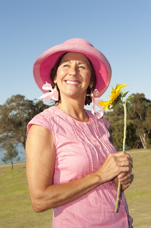 Portrait of sexy mature woman wearing pink hat and sunflower, isolated with park, lake, river and blue sky as background and copy space.の写真素材