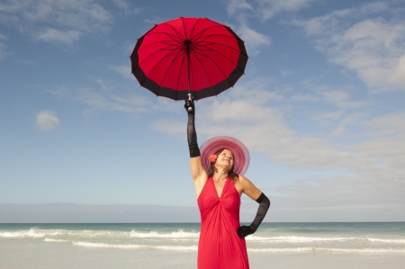 Attractive looking lady posing happy smiling in elegant red dress, black gloves and umbrella at beach, isolated with ocean and blue sky as background and copy space.の写真素材