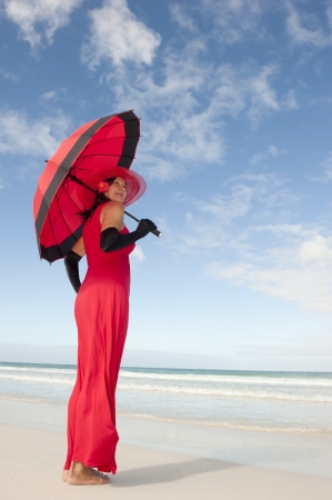 Beautiful looking lady posing happy smiling in elegant red dress, black gloves and umbrella at beach, isolated with ocean and blue sky as background and copy space.の写真素材