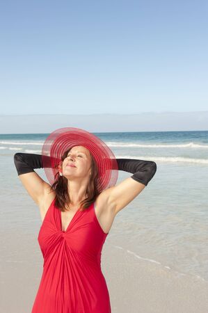 Beautiful looking lady posing happy smiling in elegant red dress and long black gloves at beach, isolated with ocean and blue sky as background and copy space.の写真素材
