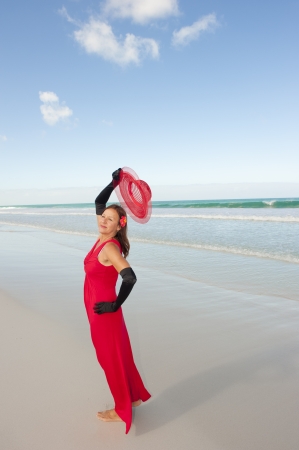 Beautiful looking lady posing happy smiling in elegant red dress and long black gloves at beach, isolated with ocean and blue sky as background and copy space.の写真素材