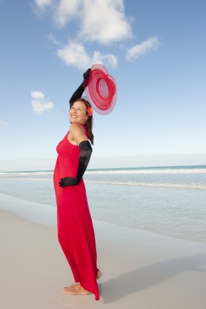 Beautiful looking lady posing happy smiling in elegant red dress and long black gloves at beach, isolated with ocean and blue sky as background and copy space.の写真素材