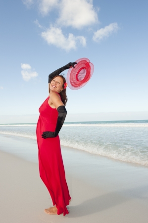 Beautiful looking lady posing happy smiling in elegant red dress and long black gloves at beach, isolated with ocean and blue sky as background and copy space.の写真素材