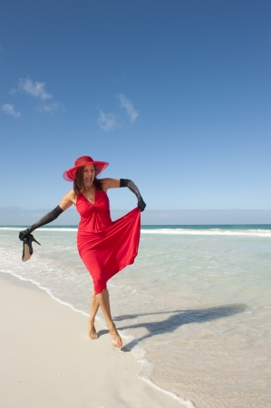 Sexy looking lady posing happy smiling in elegant red dress, long black gloves and high heels at beach, isolated with ocean and blue sky as background and copy space.の写真素材