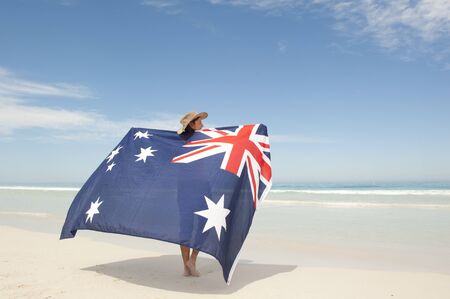Attractive mature woman wearing akubra hat and with Australian flag around shoulder standing at tropical Australian beach, isolated with ocean and blue sky as background and copy space.の写真素材