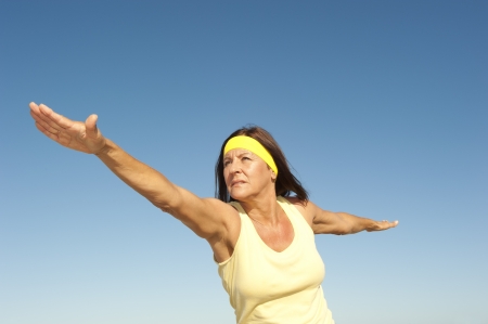Portrait attractive fit and healthy mature woman relaxed exercising outdoor with arms spread out balancing, isolated with blue sky as background and copy space.の写真素材