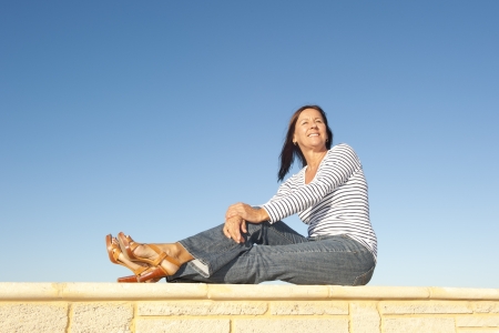 Portrait attractive mature woman sitting on a sunny day relaxed and happy on limestone wall, isolated with clear blue sky as background and copy space.の写真素材