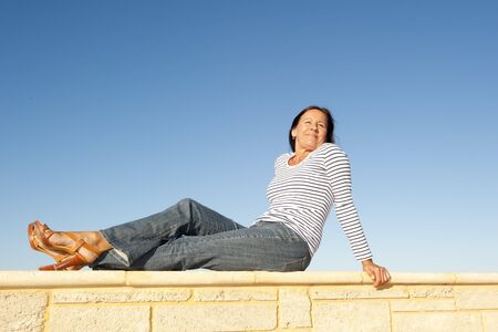 Portrait pretty mature woman sitting on a sunny day relaxed and happy on limestone wall, isolated with clear blue sky as background and copy space.の写真素材