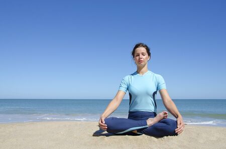 Beautiful sexy young girl sitting in cross-legged yoga lotus pose at a remote tropical beach. Clear blue sky with copy spaceの写真素材