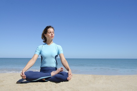 Beautiful sexy young girl sitting in cross-legged yoga lotus pose at a remote tropical beach. Clear blue sky with copy spaceの写真素材