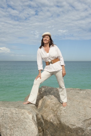 Portrait beautiful mature woman dressed in white and standing in relaxed and confident pose on rock at coastの写真素材