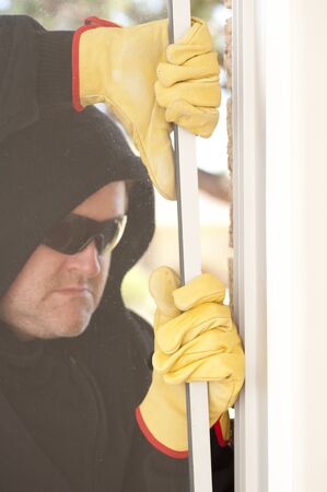 Male burglar with black hood, gloves and sunglasses trying to break in residential house, home,  through window, with copy space.の写真素材